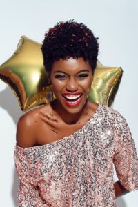 photo of laughing woman with confetti on her hair and shoulders posing in front of white background while holding golden star balloons behind her back